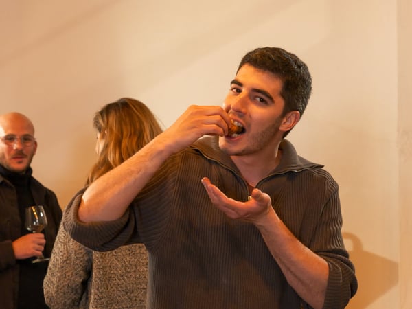 A young man in a grey ribbed sweater is captured mid-bite while eating a small snack at a social gathering, with other guests visible in the background.