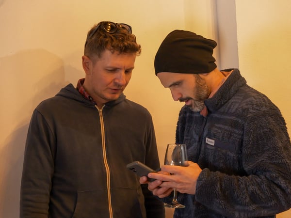 Two men are standing indoors, looking down at a smartphone that one of them is holding. One man wears a blue hoodie and sunglasses on his head, while the other wears a black beanie and a blue fleece jacket and holds a wine glass.