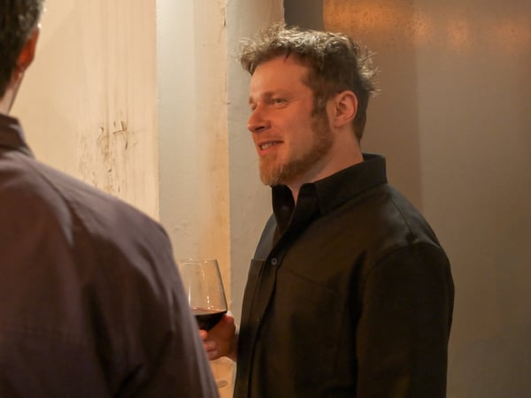 A man with a beard and messy hair holds a glass of red wine while talking to another person whose back is to the camera.