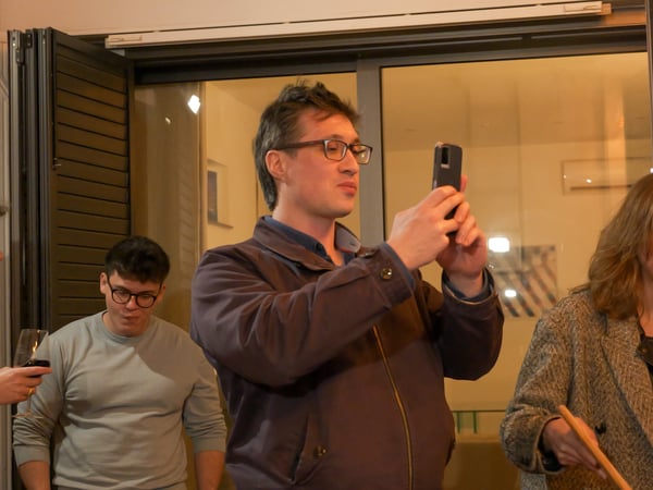 A man in a purple jacket and glasses is taking a photo with his smartphone during a social gathering. Several other people are visible in the background, including a man with a wine glass and a woman looking down.