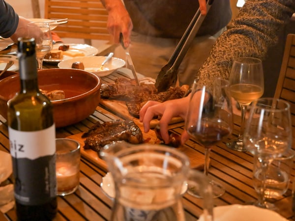A group of people are gathered around a wooden table outdoors, sharing a meal that includes grilled meat and wine.