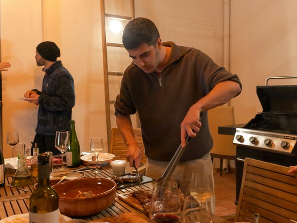 A man in a brown sweater is carving grilled meat on a wooden table during a social gathering, with a barbecue grill and another person visible in the background.