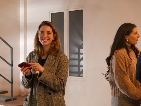 A woman in a grey coat smiles warmly at the camera while holding a red smartphone, with another woman partially visible in profile on the right.