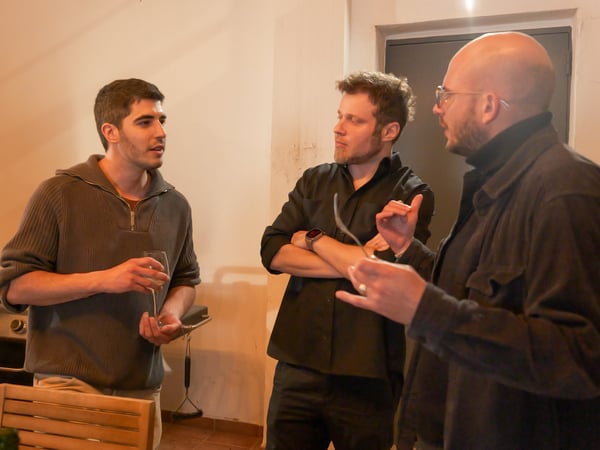 Three men are standing and engaged in a conversation in an indoor or semi-outdoor setting. One man on the left holds a wine glass while the others listen and gesture.
