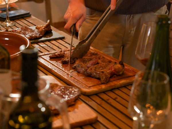 A person is using a knife and tongs to slice a cooked steak on a wooden board at a dinner table with wine bottles and glasses.