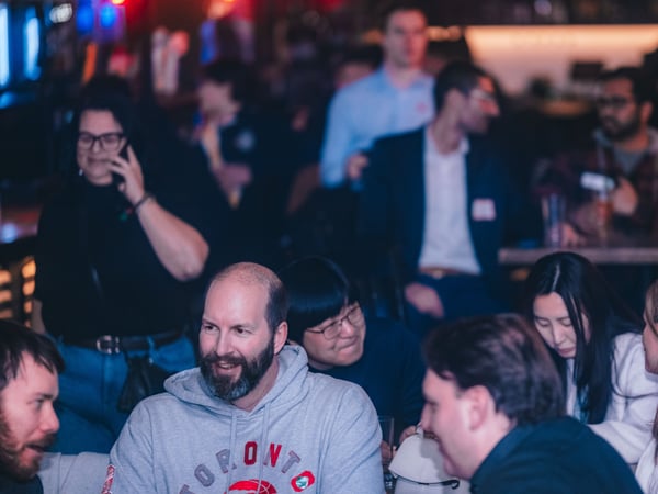 A group of people are gathered in a dimly lit indoor social setting, likely a bar or event space, engaged in various conversations.