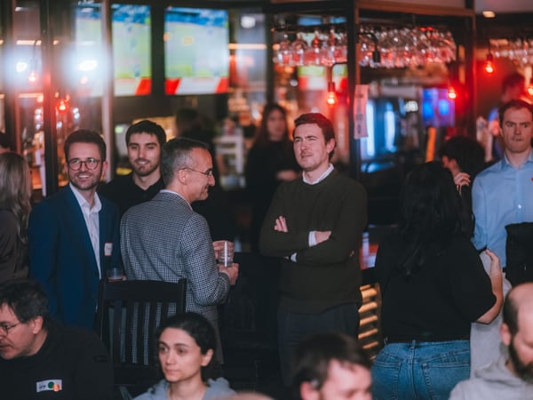 A group of people are socializing and networking in a dimly lit bar or restaurant setting with warm ambient lighting.