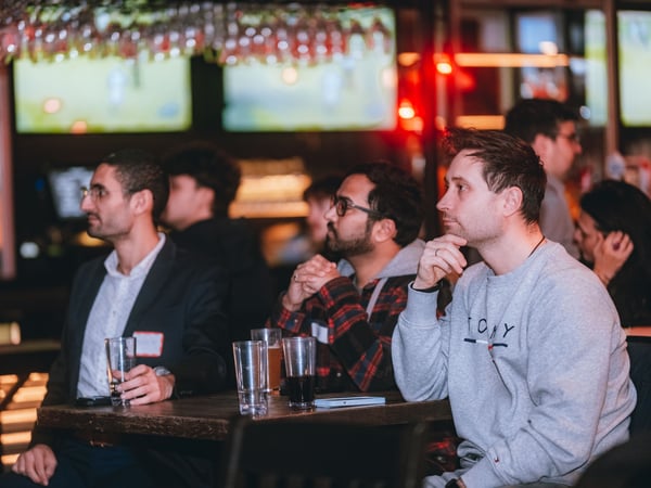 A group of people are seated at a wooden table in a bar or pub, focused on something out of frame, with drinks in front of them.