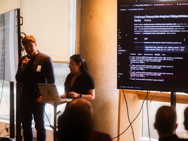 A man and a woman are giving a technical presentation, with a large screen behind them displaying code and a terminal window.