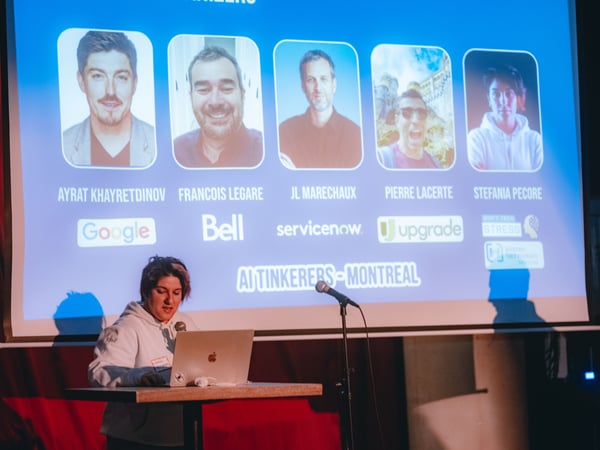 A woman is giving a presentation at an event, standing at a podium with a laptop in front of a large projection screen displaying headshots and company logos.