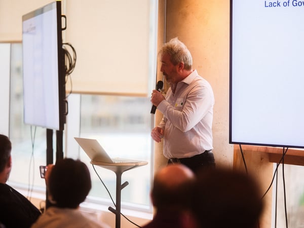 A man is giving a presentation in an indoor setting, holding a microphone and standing near a laptop and large display screens.