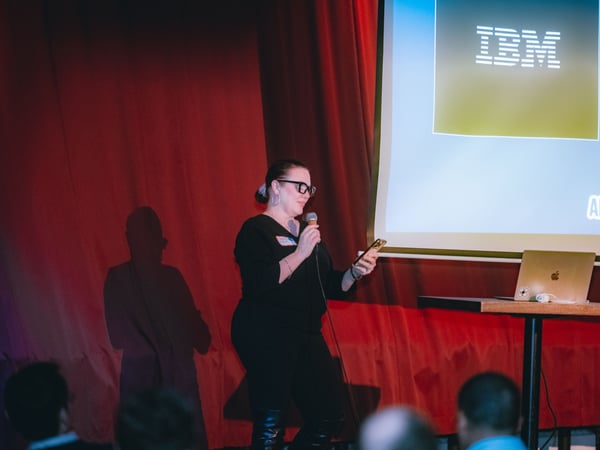 A woman stands on a stage with red curtains, holding a microphone and looking at her phone while giving a presentation. A large screen behind her displays the IBM logo and the text 'AI TINKERERS'.
