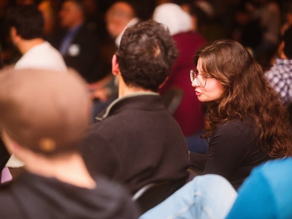 A woman with long brown hair and glasses is captured in profile while talking to someone in a crowded indoor audience.