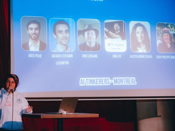 A person is giving a presentation on a stage, standing next to a laptop and speaking into a microphone. Behind them, a large screen displays the title 'AI TINKERERS - MONTREAL' along with photos and names of six individuals.