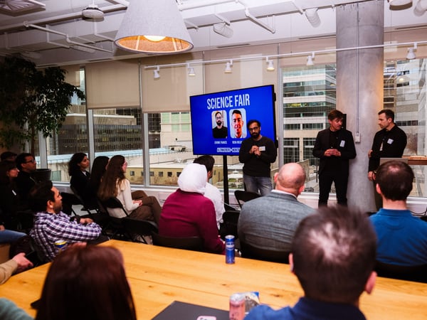 A group of people are gathered in a modern office space for a presentation titled 'Science Fair'. Three men stand at the front near a large screen, while an audience sits in chairs and at a long wooden table facing them.