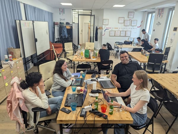 A candid photograph capturing a group of people collaborating and working on laptops around wooden tables in a bright, casual office or co-working space. The foreground shows four individuals smiling and engaged in discussion, with several others working in the background.