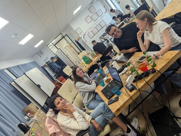 A candid photograph captures a group of people working and socializing around long wooden desks in a brightly lit, modern office environment. Several individuals in the foreground are smiling towards the camera, surrounded by laptops, power strips, and various drinks.
