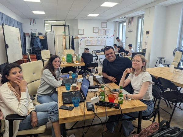 Four colleagues are seated at a wooden table, smiling directly at the camera while working on laptops in a bright, casual office or co-working space. Other people are visible working at desks further back in the room.
