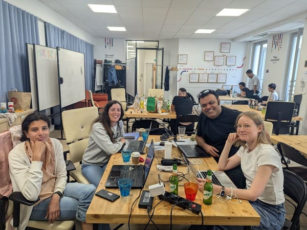 A candid photograph captures a diverse group of colleagues sitting at a wooden table, working on laptops and socializing in a brightly lit, casual office or co-working space. Four individuals in the foreground are smiling directly at the camera, while other workers are visible in the background.