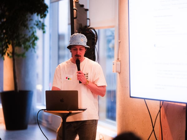 A man wearing a bucket hat and a white t-shirt stands at a small table with a laptop, speaking into a microphone during a presentation.