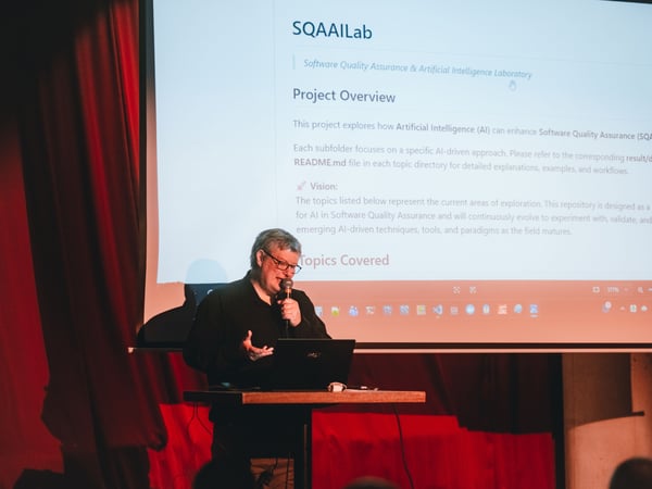 A man is giving a presentation on a stage, standing at a podium with a laptop and a microphone in front of a large projection screen displaying information about a software laboratory.