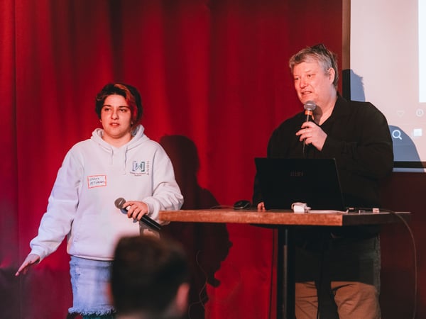 Two individuals are on a stage with a red curtain background, one holding a microphone and the other speaking into one while standing at a high table with a laptop.