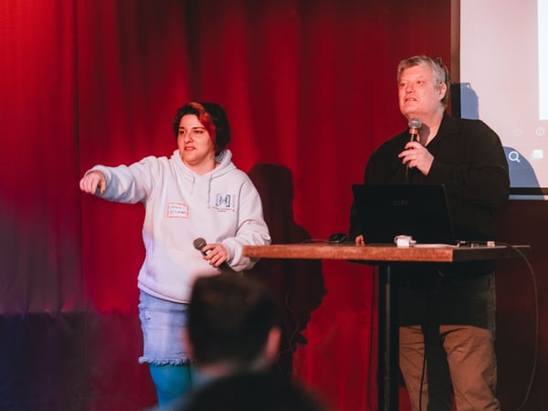 Two people are on a stage with red curtains; one in a white hoodie is pointing while holding a microphone, and another is standing behind a laptop also holding a microphone.