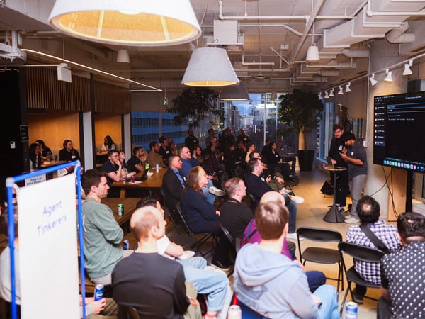 A candid indoor photograph of a group of people gathered around a table with laptops in a modern space with industrial ceiling details.