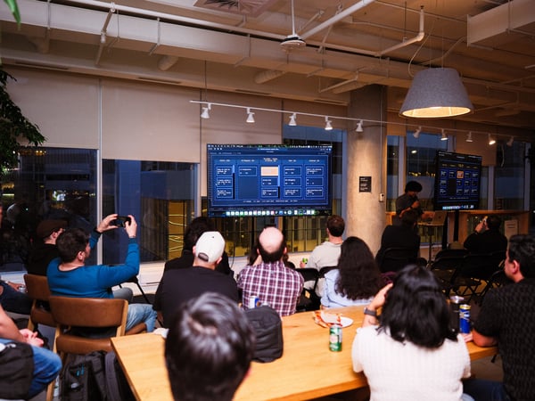 A group of people are gathered in a modern office space, watching a presentation displayed on a large screen.