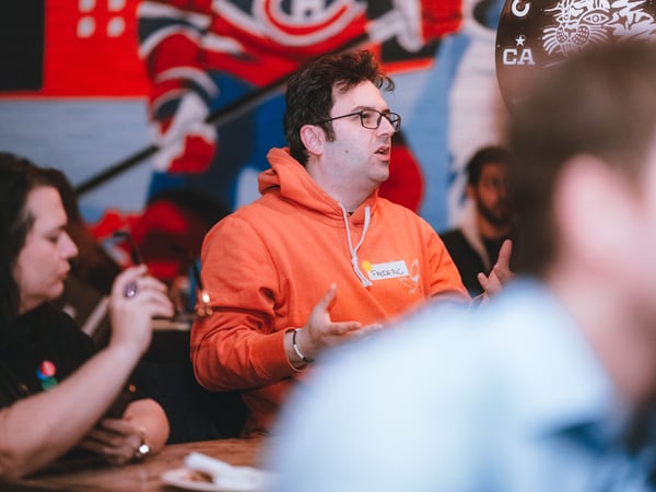A man wearing an orange hoodie and glasses is gesturing while speaking during a social gathering in a room with a sports mural.