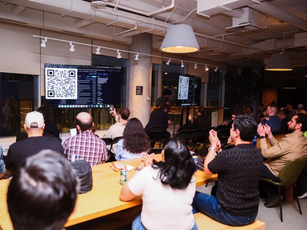 A group of people are gathered in a modern office space for a presentation, with a large screen displaying a QR code and technical information.