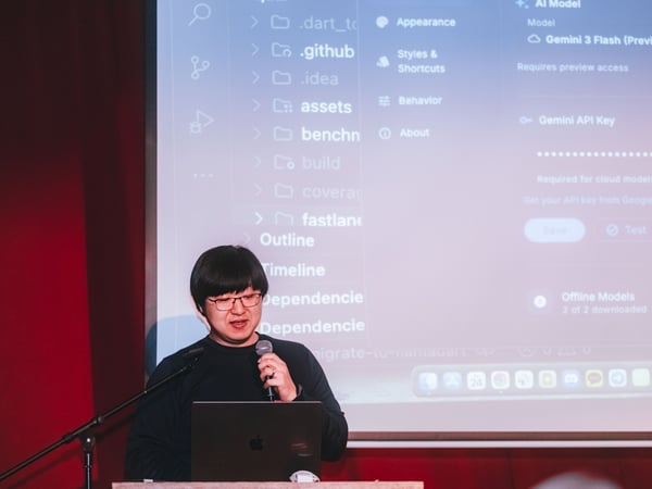 A man with glasses is giving a presentation, holding a microphone and standing behind a laptop with a large screen showing a software interface in the background.