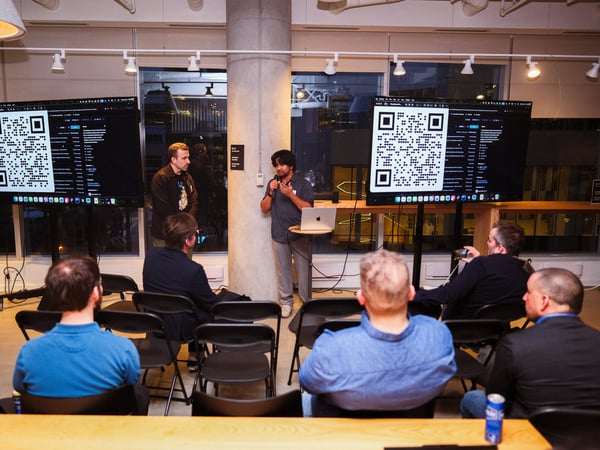A man stands between two large screens displaying QR codes and code snippets while giving a presentation to a seated audience in a modern office or event space.