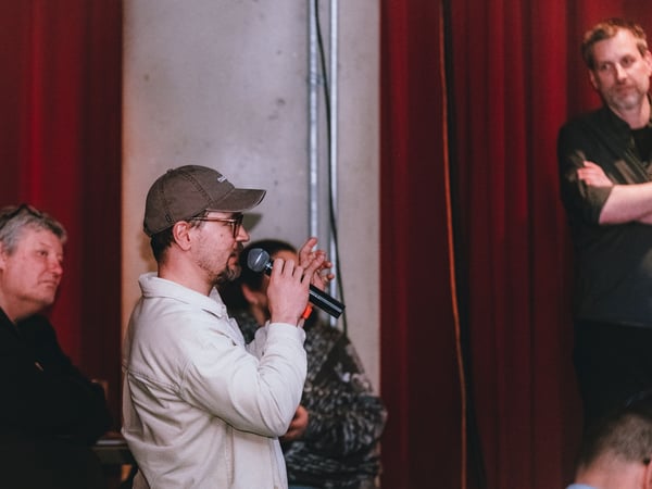 A man wearing a baseball cap and glasses speaks into a microphone during an indoor event, with other attendees and red curtains in the background.