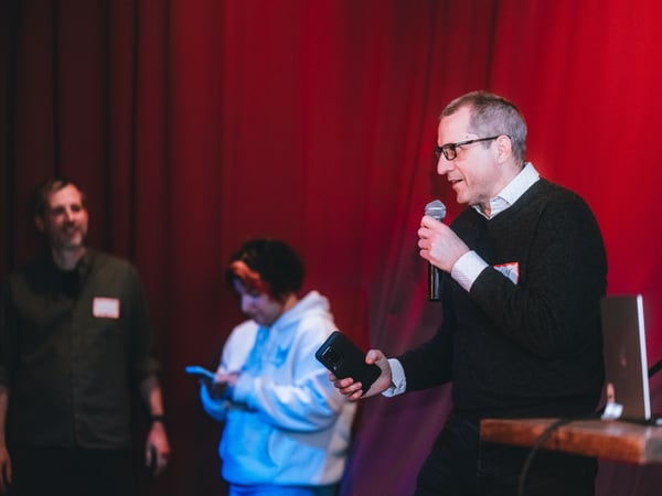 A man in a black sweater and glasses speaks into a microphone on a stage with a red curtain backdrop, while two other people stand in the background.