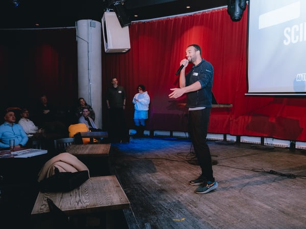 A man stands on a stage giving a presentation with a microphone in front of a red curtain and a projection screen. An audience is seated at wooden tables in the foreground of the dimly lit venue.