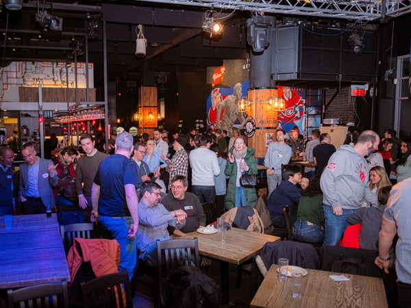 A crowded indoor social gathering at a bar or restaurant, featuring many people standing and sitting while talking. The venue has high ceilings with stage lighting and a large mural on the back wall.