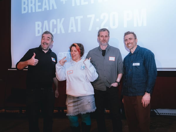 Four people, three men and one woman, pose for a group photo in front of a projection screen during an event break.