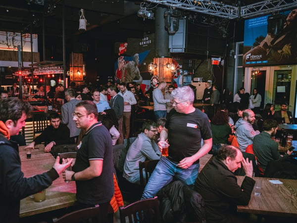 A wide-angle photograph of a crowded indoor social event at a bar or pub, featuring many people standing and sitting while talking and drinking.