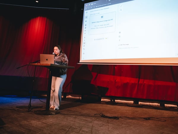 A woman is giving a presentation on a stage, standing behind a podium with a laptop while a large screen behind her displays a web application interface.