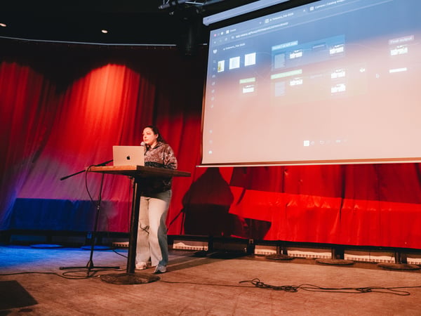 A woman stands at a podium on a stage, giving a presentation with a large screen behind her displaying a software interface. The stage is lit with red and blue lights, and a laptop is open on the podium.