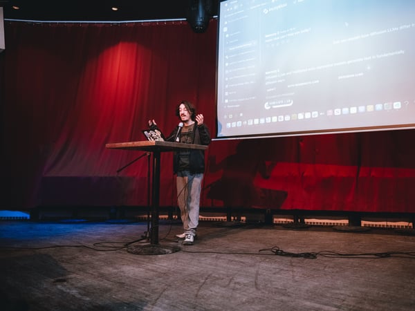 A man is giving a presentation on a stage, standing at a high table with a laptop in front of a large projection screen displaying technical information about LLMs.
