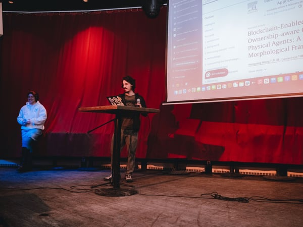 A man stands at a podium on a stage, giving a presentation with a large screen behind him displaying a research paper about blockchain and cyber-physical agents. A woman stands to the side, watching the presentation against a backdrop of red curtains.