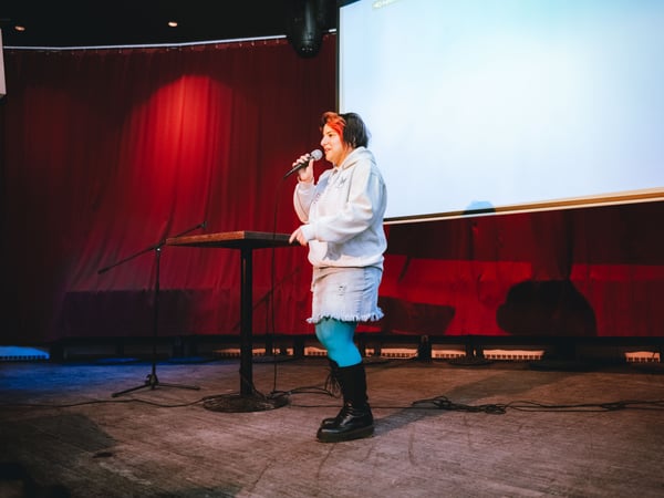 A person with vibrant hair stands on a stage, speaking into a microphone in front of a large projection screen and a red curtain.