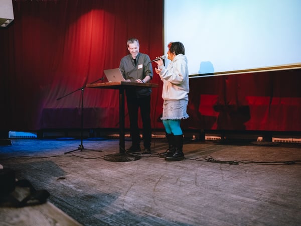 Two individuals are on a stage during an event; one person is using a laptop at a high table while the other speaks into a microphone.
