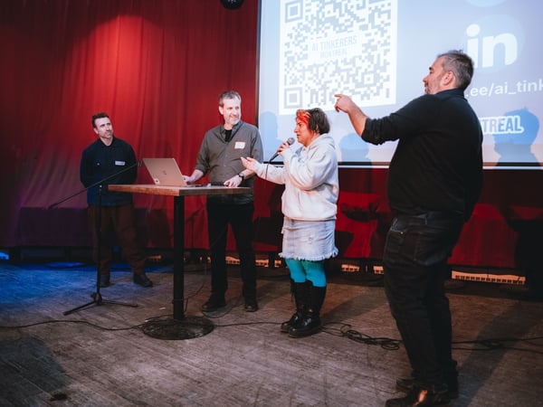 A group of people are on a stage during a presentation event. One person is speaking into a microphone while another stands at a laptop on a high table, with a large projection screen in the background.