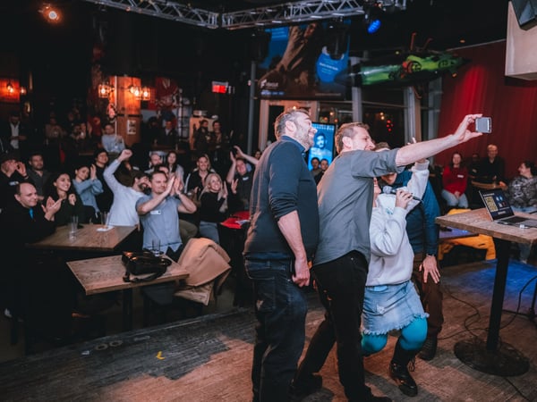 A group of people on stage take a selfie with a smartphone while an audience in the background cheers and claps at an indoor event.