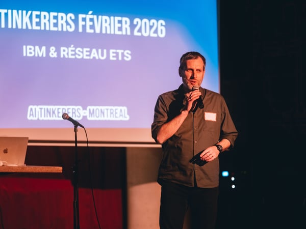 A man is speaking into a microphone on a stage in front of a projection screen for an AI Tinkerers event in Montreal.