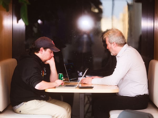 Two men are sitting at a table in a booth, focused on their laptops in an indoor setting.