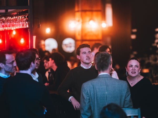 A group of people socializing in a dimly lit, warm bar or lounge setting with soft, glowing lights in the background.